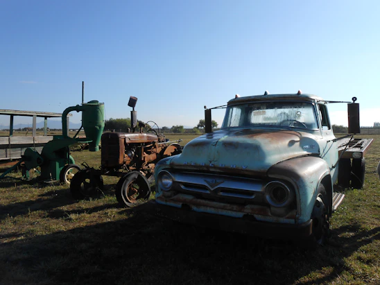 An old, rusty truck alongside an antique tractor and green farm machinery are parked in an open field under a clear blue sky. The vehicles show signs of wear and age, with patches of rust and faded paint. The scene appears to be set in a rural area with distant trees and flat terrain visible in the background.