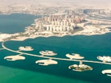 An aerial view of a coastal city with numerous high-rise buildings surrounded by water and man-made islands. The area features intricate land formations and several bridges connecting the islands. The cityscape is densely packed, showcasing urban development against a backdrop of desert.