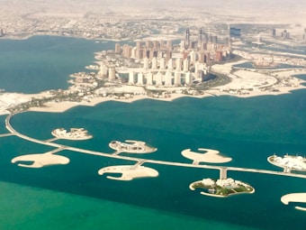 An aerial view of a coastal city with numerous high-rise buildings surrounded by water and man-made islands. The area features intricate land formations and several bridges connecting the islands. The cityscape is densely packed, showcasing urban development against a backdrop of desert.