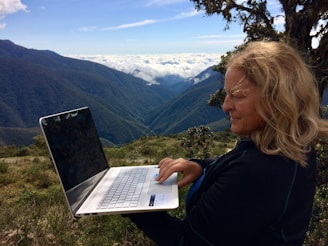 A person is using a laptop in a scenic mountainous landscape with sprawling greenery and a backdrop of clouds. The mountains stretch into the distance under a clear blue sky.