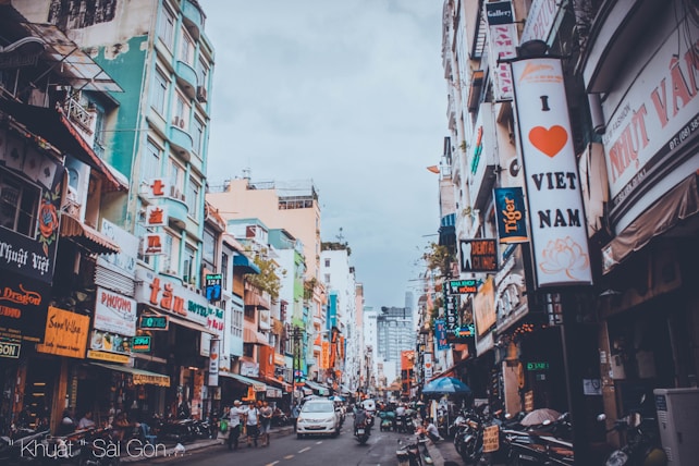A bustling urban street lined with colorful multi-story buildings adorned with various signs and advertisements. Brightly lit shopfronts and a mix of traditional and modern architectural styles are visible. People and motorbikes fill the street, contributing to the lively atmosphere.