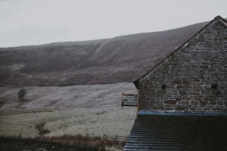 A rustic stone building stands in front of a smooth, grassy hillside. The structure features an exposed brick wall and a well-weathered roof. A sign on the building reads 'Dalehead Bunkhouse'. The surrounding landscape appears serene, with muted colors and a slightly overcast sky.