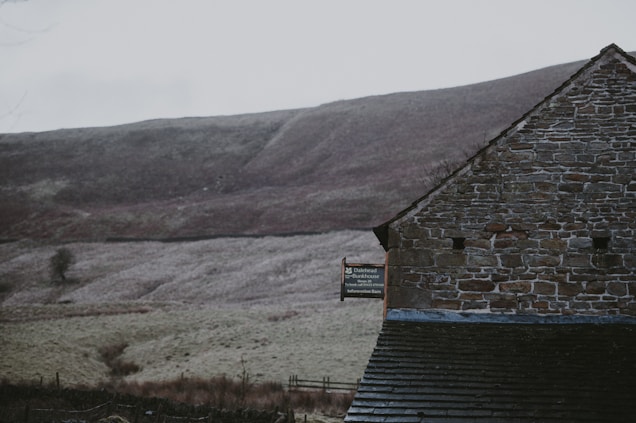 A rustic stone building stands in front of a smooth, grassy hillside. The structure features an exposed brick wall and a well-weathered roof. A sign on the building reads 'Dalehead Bunkhouse'. The surrounding landscape appears serene, with muted colors and a slightly overcast sky.