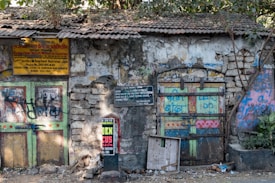 A rustic, deteriorating building facade features two colorful wooden doors, covered in graffiti and surrounded by crumbling stone walls. Signs with Hindi script hang above the doors, and an advertisement is posted on a nearby utility pole. The roof is made of old tiles and small plants can be seen growing near the structure.