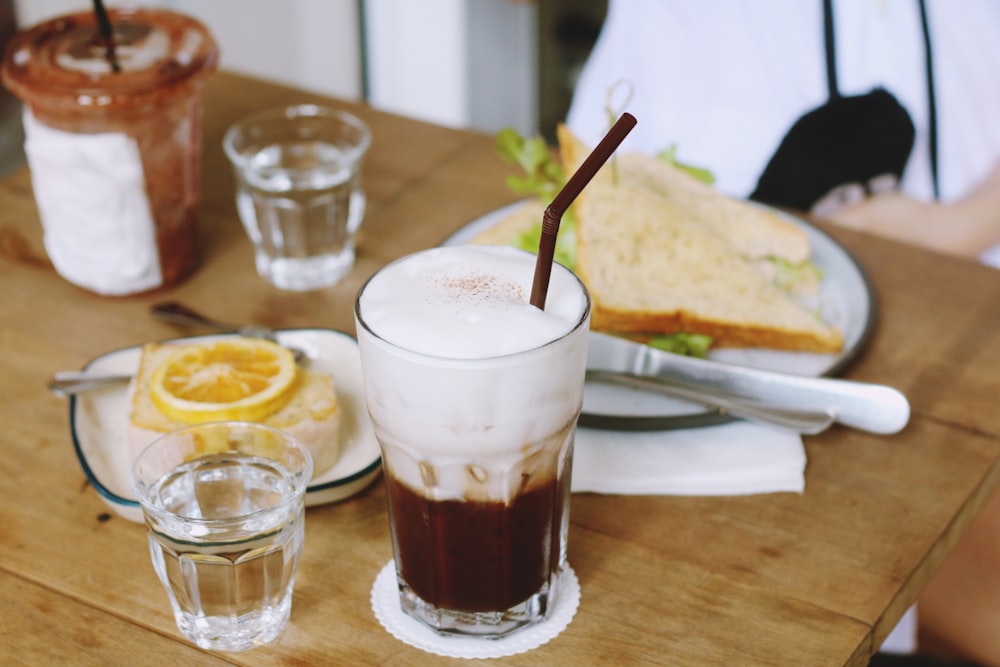 clear drinking glass filled with brown liquid in front of plate sandwich on table