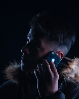 A person is holding a smartphone to their ear, looking contemplatively upward. The lighting is dark, with highlights illuminating part of their face and the fur trim of their jacket.