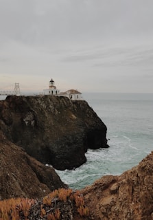 A coastal landscape featuring a lighthouse perched on a rocky cliff overlooking the ocean. Waves crash against the base of the cliffs, and a rugged pathway leads to the lighthouse. The sky is overcast, adding a moody backdrop to the scene. Sparse vegetation clings to the rock surfaces, and a small building accompanies the lighthouse.