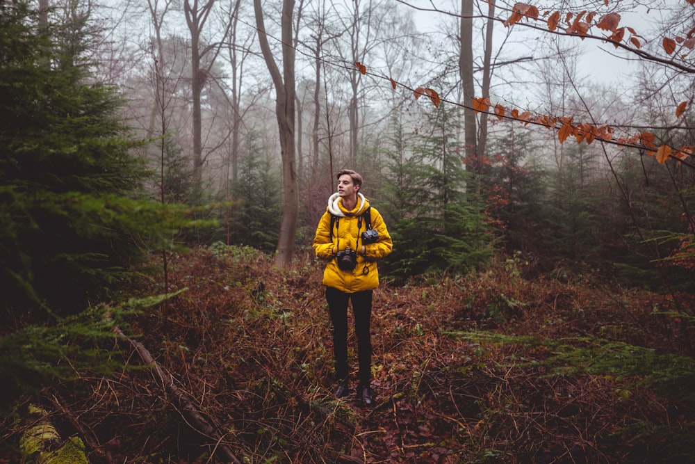 homem na jaqueta amarela segurando a câmera preta em pé em gramíneas marrons sob as árvores