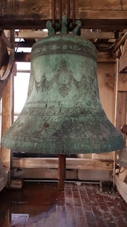 A close-up of a finely crafted cast bell with weathered patina hanging in a wooden tower frame.