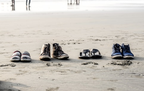 Four pairs of footwear, including casual shoes, boots, sandals, and sneakers, are neatly arranged in a row on the sandy beach. In the background, the shoreline and a few figures can be seen blurred in the distance.