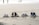 Four pairs of footwear, including casual shoes, boots, sandals, and sneakers, are neatly arranged in a row on the sandy beach. In the background, the shoreline and a few figures can be seen blurred in the distance.