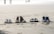 Four pairs of footwear, including casual shoes, boots, sandals, and sneakers, are neatly arranged in a row on the sandy beach. In the background, the shoreline and a few figures can be seen blurred in the distance.