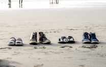 Four pairs of footwear, including casual shoes, boots, sandals, and sneakers, are neatly arranged in a row on the sandy beach. In the background, the shoreline and a few figures can be seen blurred in the distance.