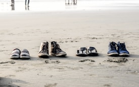 Four pairs of footwear, including casual shoes, boots, sandals, and sneakers, are neatly arranged in a row on the sandy beach. In the background, the shoreline and a few figures can be seen blurred in the distance.