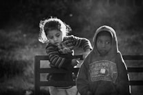 Two children are sitting on a bench. The child on the left is wearing a striped sweater and appears to be leaning towards the child on the right, who is wearing a hoodie. The background is blurred, giving prominence to the children.