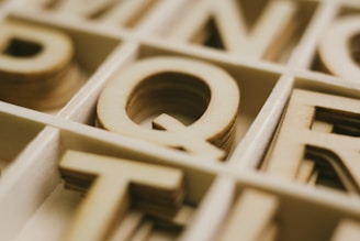 Wooden letters arranged in a grid, with the letter Q prominently centered. The letters have a natural wooden texture and appear to be part of a type set.