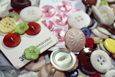 A variety of buttons in different sizes, colors, and materials are scattered and some are attached to cards. Patterns include striped, solid, and textured designs. The buttons are primarily circular with different numbers of holes, and some cards display branding and price details.