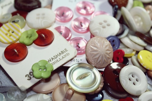 A variety of buttons in different sizes, colors, and materials are scattered and some are attached to cards. Patterns include striped, solid, and textured designs. The buttons are primarily circular with different numbers of holes, and some cards display branding and price details.