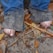 Bare feet covered in dirt and sand are partially visible under frayed blue jeans. The ground is scattered with dried leaves and twigs.