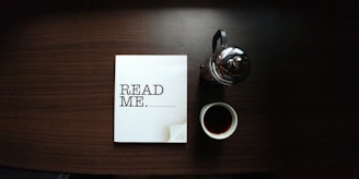 Papirbog med teksten: Read Me. Ved siden af en stempelkande og et kaffekrus på et bord.