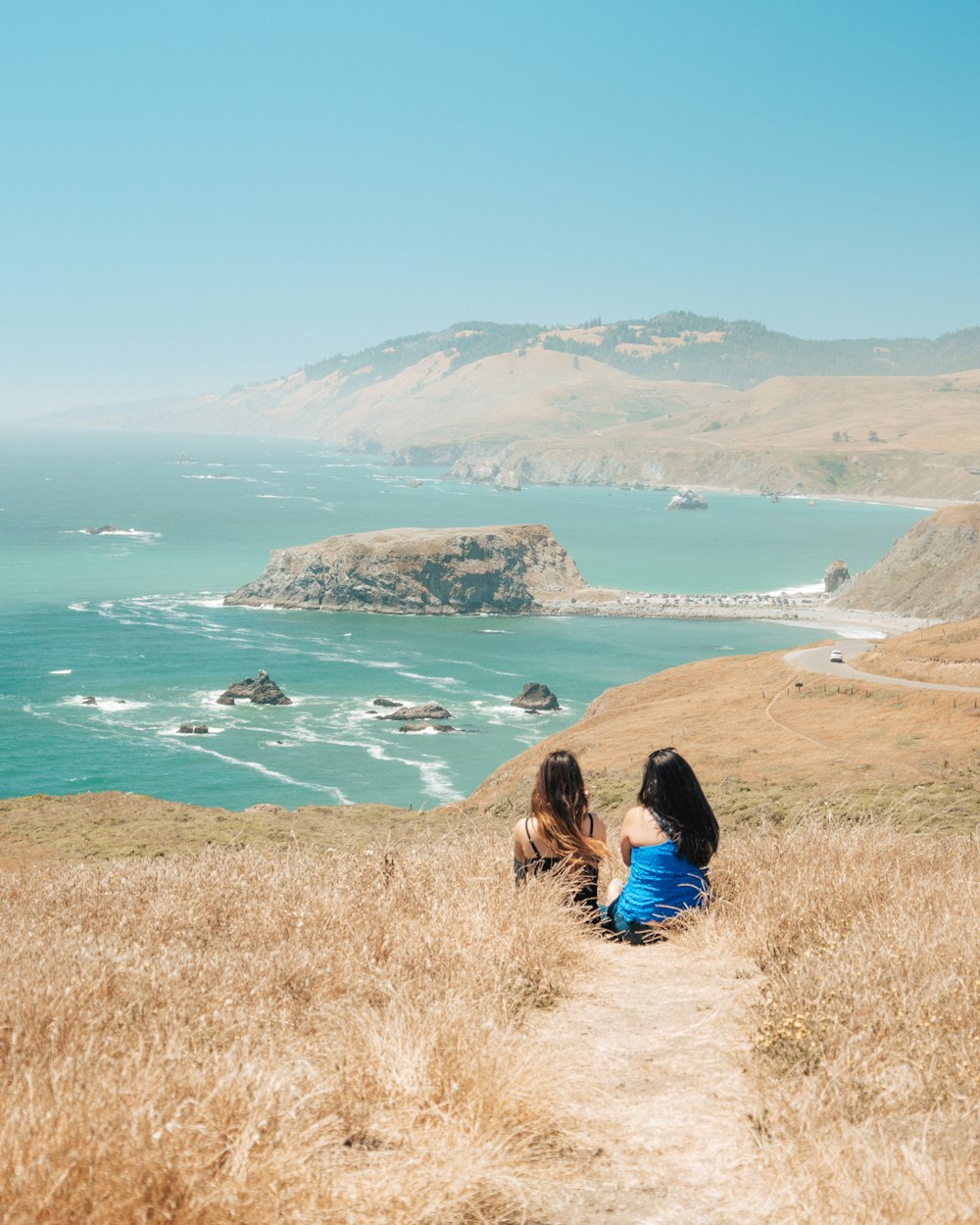 two women sitting on brown grass facing mountain and sea water