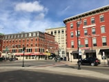 An urban street corner featuring several multi-story red brick buildings. These buildings house various businesses, including a healthcare office and a donut shop. The sky is partly cloudy, and there are some vehicles and pedestrian traffic in the area. Street signs and traffic lights are visible, adding to the typical cityscape ambiance.