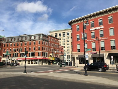 An urban street corner featuring several multi-story red brick buildings. These buildings house various businesses, including a healthcare office and a donut shop. The sky is partly cloudy, and there are some vehicles and pedestrian traffic in the area. Street signs and traffic lights are visible, adding to the typical cityscape ambiance.