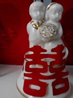 A white ceramic figurine depicting two children holding a gold-trimmed item. Red Chinese characters, likely for double happiness, enhance the decorative piece. The background is dominated by a rich red color, aligning with traditional festive themes.
