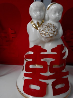 A white ceramic figurine depicting two children holding a gold-trimmed item. Red Chinese characters, likely for double happiness, enhance the decorative piece. The background is dominated by a rich red color, aligning with traditional festive themes.