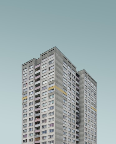 A tall, rectangular residential tower with numerous windows and balconies, featuring a minimalist design against a pale sky background.