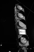 A black and white photograph of a traffic light with four lights stacked vertically. Each light is encased in a round hood, with the bottom section labeled 'M252'. The traffic light is against a dark background with faint lines and distant small lights visible.