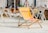 A wooden deck illustrates a relaxed setting with multiple folded metal chairs and sun loungers, hinting at a beach or poolside location. The center features an orange folding chair branded 'Schöfferhofer Grapefruit,' adorned with a grapefruit image. In the background stands a play area with equipment like slides and climbing structures, surrounded by children and some adults sunbathing.
