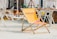 A wooden deck illustrates a relaxed setting with multiple folded metal chairs and sun loungers, hinting at a beach or poolside location. The center features an orange folding chair branded 'Schöfferhofer Grapefruit,' adorned with a grapefruit image. In the background stands a play area with equipment like slides and climbing structures, surrounded by children and some adults sunbathing.
