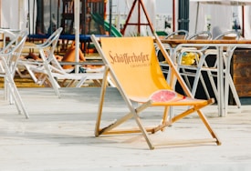 A wooden deck illustrates a relaxed setting with multiple folded metal chairs and sun loungers, hinting at a beach or poolside location. The center features an orange folding chair branded 'Sch&ouml;fferhofer Grapefruit,' adorned with a grapefruit image. In the background stands a play area with equipment like slides and climbing structures, surrounded by children and some adults sunbathing.