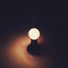 An illuminated light bulb stands upright with a dark background. The light is casting a warm glow on the surface it is placed upon, creating a serene and introspective atmosphere.