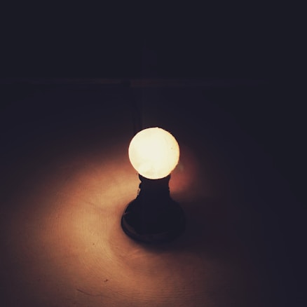 An illuminated light bulb stands upright with a dark background. The light is casting a warm glow on the surface it is placed upon, creating a serene and introspective atmosphere.