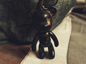 A small, stylized bear figurine with a glossy finish stands upright. The bear is black with gold lettering and designs, including the words 'SPACE & PEACE'. The background consists of soft, muted fabrics, adding a cozy atmosphere.