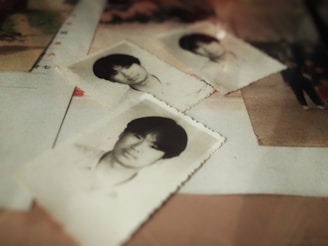 A collection of black and white photographs depicting a young man, scattered on a surface. The images appear aged, with slightly curled edges, conveying a sense of nostalgia. Other papers or photographs are partially visible beneath.