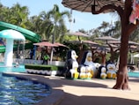 A water park setting with a swimming pool area. Several large penguin sculptures are arranged near the poolside. Palm trees and green foliage surround the area, creating a tropical atmosphere. A winding green waterslide is visible in the background. There are people enjoying the pool, and multiple large sun umbrellas provide shade.