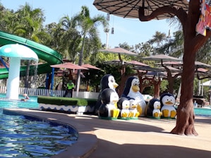A water park setting with a swimming pool area. Several large penguin sculptures are arranged near the poolside. Palm trees and green foliage surround the area, creating a tropical atmosphere. A winding green waterslide is visible in the background. There are people enjoying the pool, and multiple large sun umbrellas provide shade.