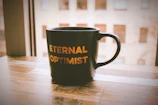 A cheerful mug with a bright, positive message resting on a wooden table beside a sunny window.