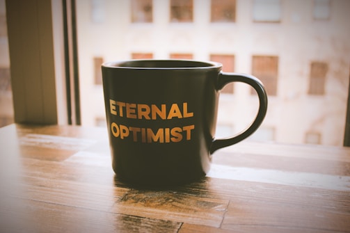 A black mug with the words 'ETERNAL OPTIMIST' printed in bold golden letters sits on a wooden table near a window. The window view shows an out-of-focus building exterior, suggesting an urban setting. Soft, warm lighting casts gentle shadows, creating a cozy atmosphere.