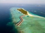 An aerial view of a tropical island with overwater bungalows extending into the turquoise sea. The island is lush with greenery and surrounded by clear blue waters. Several smaller islands can be seen in the distance, and scattered boats float near the shore.