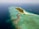 An aerial view of a tropical island with overwater bungalows extending into the turquoise sea. The island is lush with greenery and surrounded by clear blue waters. Several smaller islands can be seen in the distance, and scattered boats float near the shore.