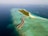 An aerial view of a tropical island with overwater bungalows extending into the turquoise sea. The island is lush with greenery and surrounded by clear blue waters. Several smaller islands can be seen in the distance, and scattered boats float near the shore.