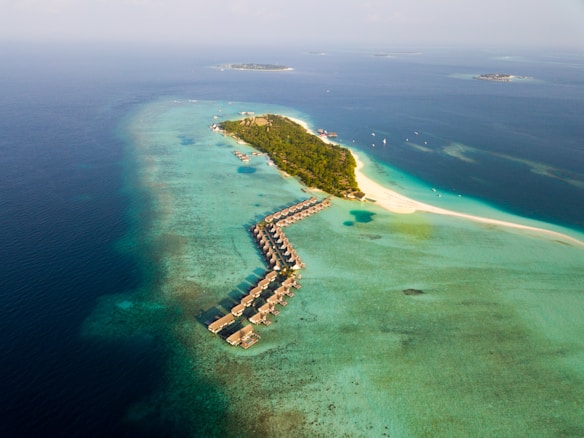 An aerial view of a tropical island with overwater bungalows extending into the turquoise sea. The island is lush with greenery and surrounded by clear blue waters. Several smaller islands can be seen in the distance, and scattered boats float near the shore.