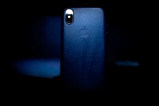 A smartphone with a sleek design is prominently featured in low light, focusing on its back side which displays the logo of a popular technology company. The surrounding is dark, creating a spotlight effect on the device.