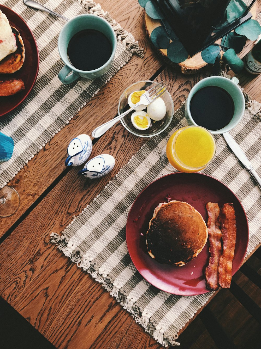gebackenes Brot auf runder roter Keramikplatte neben einem Glas Orangensaft, einer Tasse Kaffee und gekochten Eiern