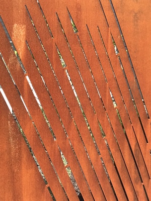 A close-up view of a rust-colored metal surface with elongated, narrow cutouts in a diagonal pattern, allowing glimpses of greenery through the openings.