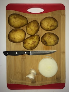 Five whole potatoes, a sliced onion, a few cloves of garlic, and a knife are arranged on a wooden cutting board with a red handle.